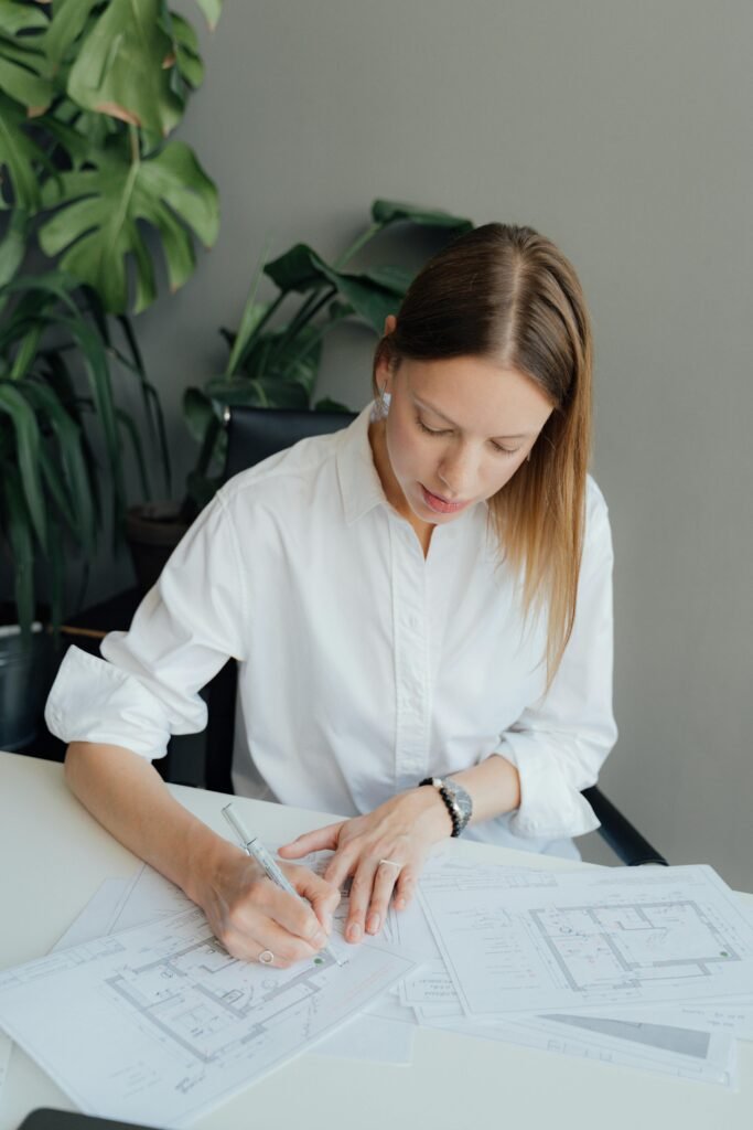 Testimonials Woman architect reviewing floor plans at a desk indoors, surrounded by plants.